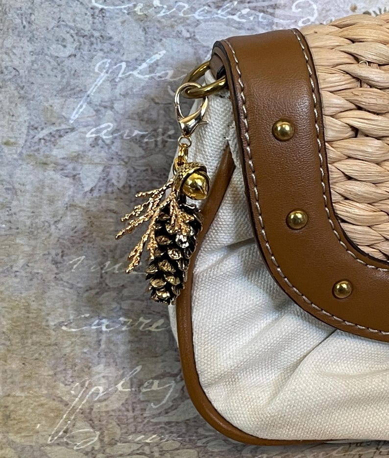 May include: A handbag with a woven straw body and brown leather trim. A gold-coloured charm shaped like a pinecone and sprig hangs from the bag's strap. The bag has a white canvas interior.