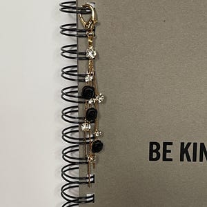 May include: A gold-colored bookmark with a decorative chain featuring black rose charms and clear crystal accents. The bookmark is attached to a gray spiral-bound notebook with the words "BE KIND" printed on the cover.