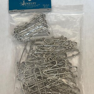 May include: A collection of silver-colored safety pins with three loops, displayed in a clear plastic bag. A single antique brass-colored safety pin with three loops is also shown. The packaging reads "The Jewelry Shoppe Findings" and "Nickel Free".