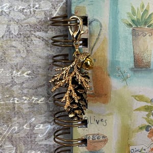 May include: A gold-coloured charm with a pinecone and acorn design, attached to a spiral-bound notebook. The notebook features a watercolour illustration of a potted plant and the word "Olives". The charm is attached with a lobster clasp.