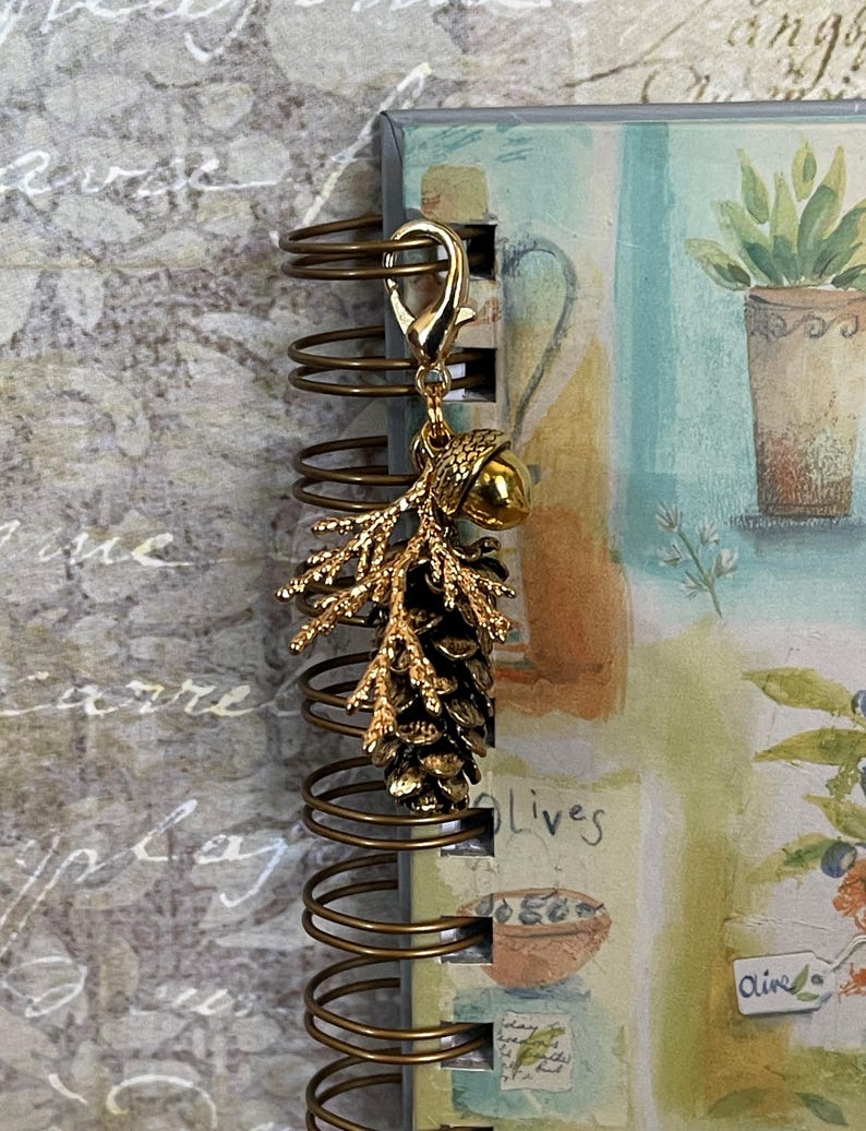 May include: A gold-coloured charm with a pinecone and acorn design, attached to a spiral-bound notebook. The notebook features a watercolour illustration of a potted plant and the word "Olives". The charm is attached with a lobster clasp.