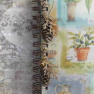 May include: Gold-coloured charm featuring a pinecone and sprig design, attached to a spiral-bound notebook. The notebook displays botanical illustrations and handwritten text. The charm is a decorative accessory.