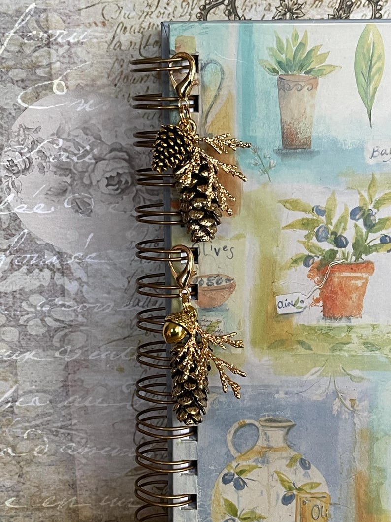 May include: Gold-coloured charm featuring a pinecone and sprig design, attached to a spiral-bound notebook. The notebook displays botanical illustrations and handwritten text. The charm is a decorative accessory.