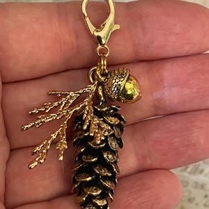 May include: A gold-coloured charm featuring a detailed pinecone, an acorn, and a sprig of leaves. The pinecone is dark brown with gold accents, and the acorn is a solid gold colour. The charm is attached to a gold-coloured clasp.
