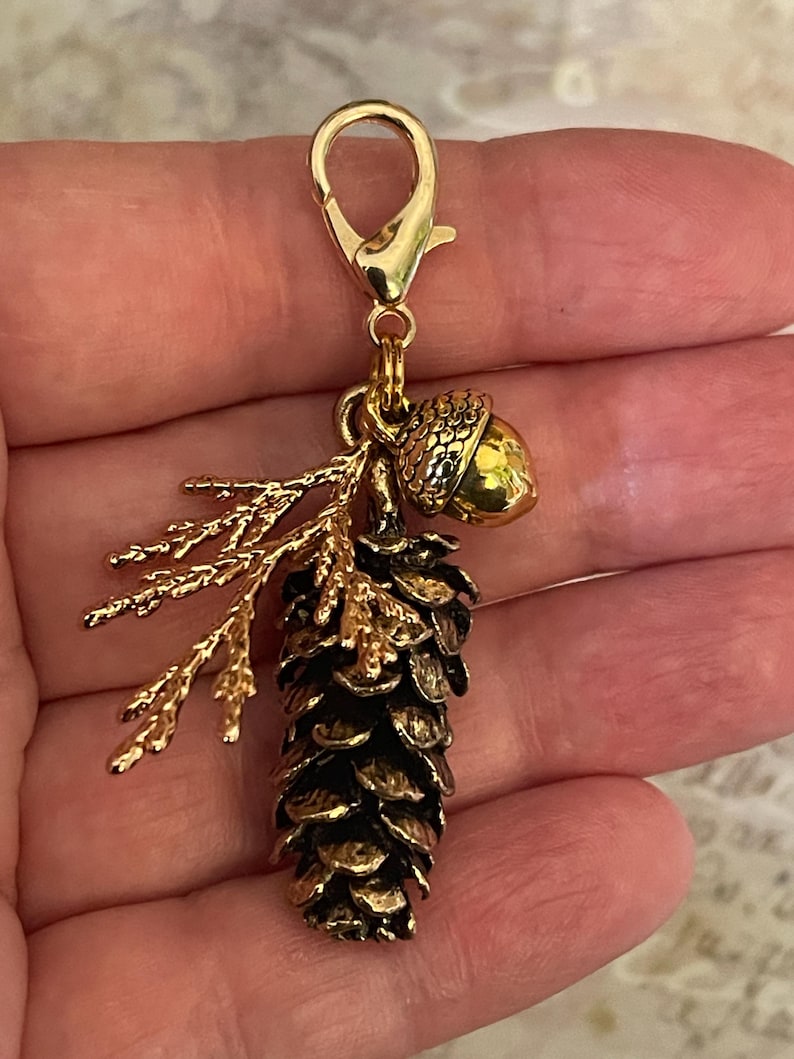 May include: A gold-coloured charm featuring a detailed pinecone, an acorn, and a sprig of leaves. The pinecone is dark brown with gold accents, and the acorn is a solid gold colour. The charm is attached to a gold-coloured clasp.