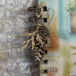May include: A gold-coloured charm with a detailed pinecone and branch design. The charm is attached to a gold-coloured clasp, suitable for a necklace or bracelet. The charm is shown next to a spiral-bound notebook.