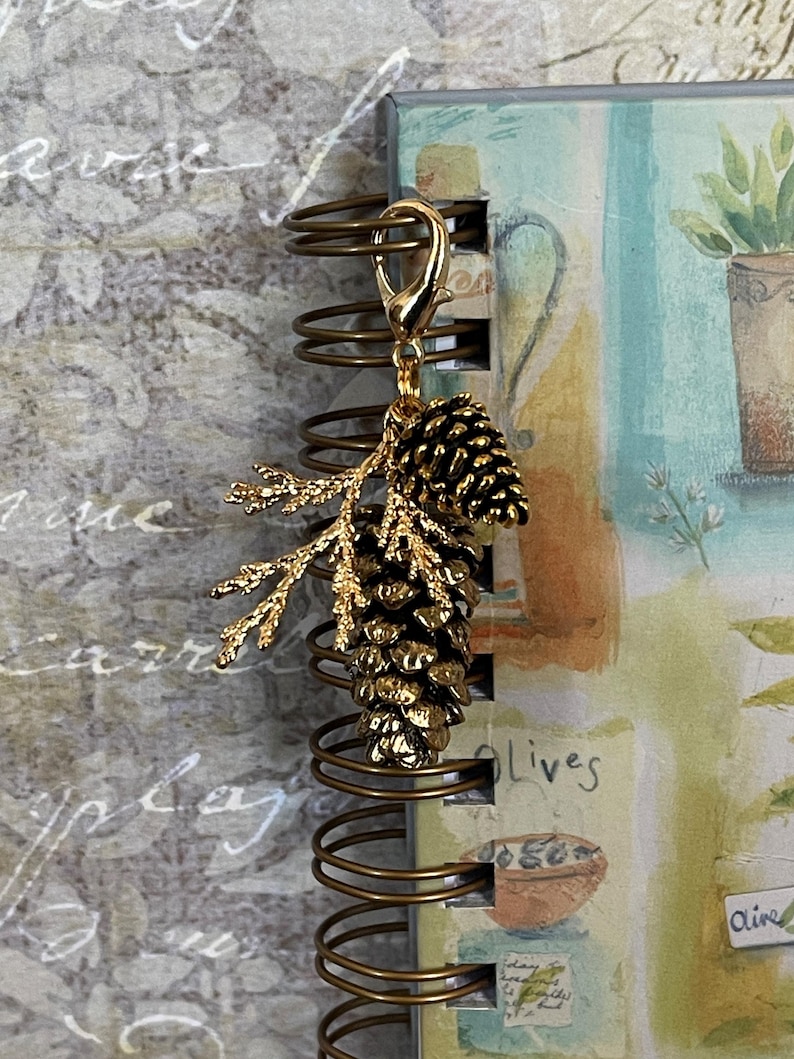 May include: A gold-coloured charm with a detailed pinecone and branch design. The charm is attached to a gold-coloured clasp, suitable for a necklace or bracelet. The charm is shown next to a spiral-bound notebook.