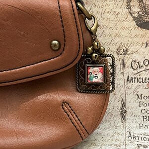 May include: A brown leather purse with a decorative bronze charm. The charm features a small square image of Santa Claus, surrounded by a decorative frame and small bells. The purse has a stitched design and a metal clasp.