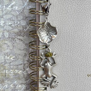 May include: A silver-toned frog charm with a green stone head hangs from a silver leaf charm. The charms are attached to a silver clasp, clipped to the spiral binding of a notebook. The notebook has a light gray cover.