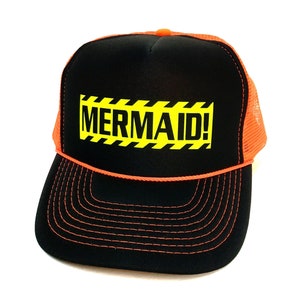 May include: A black trucker hat with an orange mesh back. The front of the hat has a yellow and black caution tape design with the text "MERMAID!" in black.