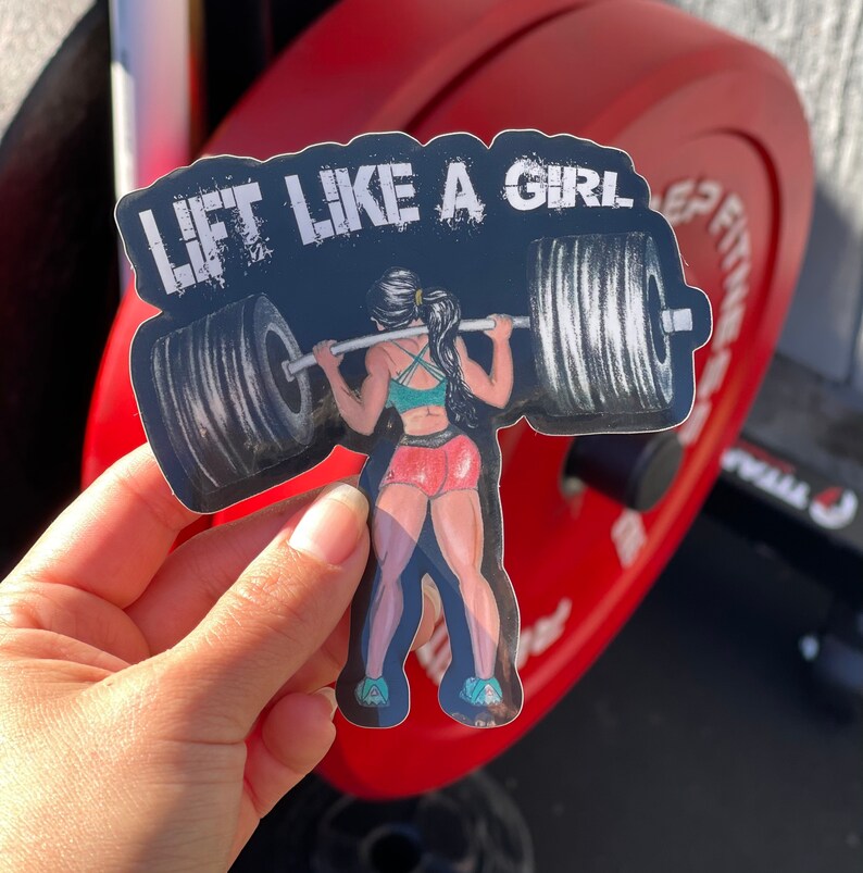 Puede incluir: Pegatina en blanco y negro con una mujer levantando pesas. La pegatina dice "Lift Like A Girl" en texto blanco.