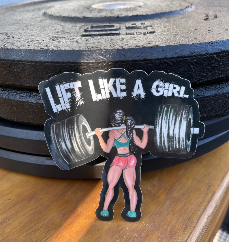 Puede incluir: Una pegatina en blanco y negro con una mujer levantando una barra. La pegatina dice "Lift Like A Girl" en letras blancas.