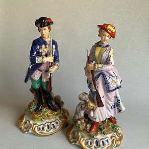 May include: A pair of porcelain figurines depicting a man and woman in hunting attire. The man is wearing a blue and pink jacket, green trousers, and a black hat. The woman is wearing a pink and blue dress with a yellow waistcoat and a red hat. Both figurines are holding rifles and are standing on ornate bases.