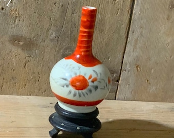 Może przedstawiać: Mały, ręcznie malowany ceramiczny wazon z białym korpusem, pomarańczowymi akcentami i czerwoną szyjką. Wazon ma kwiatowe wzory i stoi na czarnym drewnianym stojaku. Tekst "Unique pottery Shop" jest widoczny w tle.