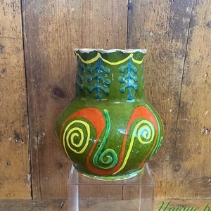 May include: A hand-painted ceramic vase with a green base, decorated with orange and yellow spiral designs. The top has a scalloped rim and blue and green leaf-like patterns. The vase is displayed on a clear stand against a wooden background.