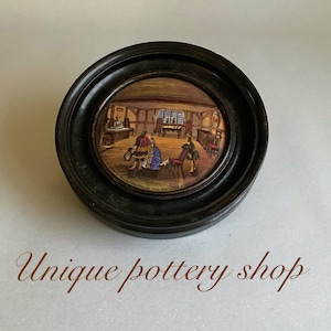 Puede incluir: Una imagen redonda enmarcada en madera de una pequeña pintura detallada del interior de una tienda de cerámica. La pintura muestra una tienda con un mostrador, estantes y personas comprando. El marco es negro y tiene un borde elevado.