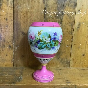 May include: A vintage porcelain vase with a pink and white floral design. The vase has a round body and a pedestal base. The floral design features pink, white, and purple flowers with green leaves.