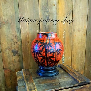 Op de afbeelding: Een rode en zwarte keramische vaas met een bamboe ontwerp. De vaas staat op een houten kist. De tekst "Unique Pottery Shop" is zichtbaar op de achtergrond.