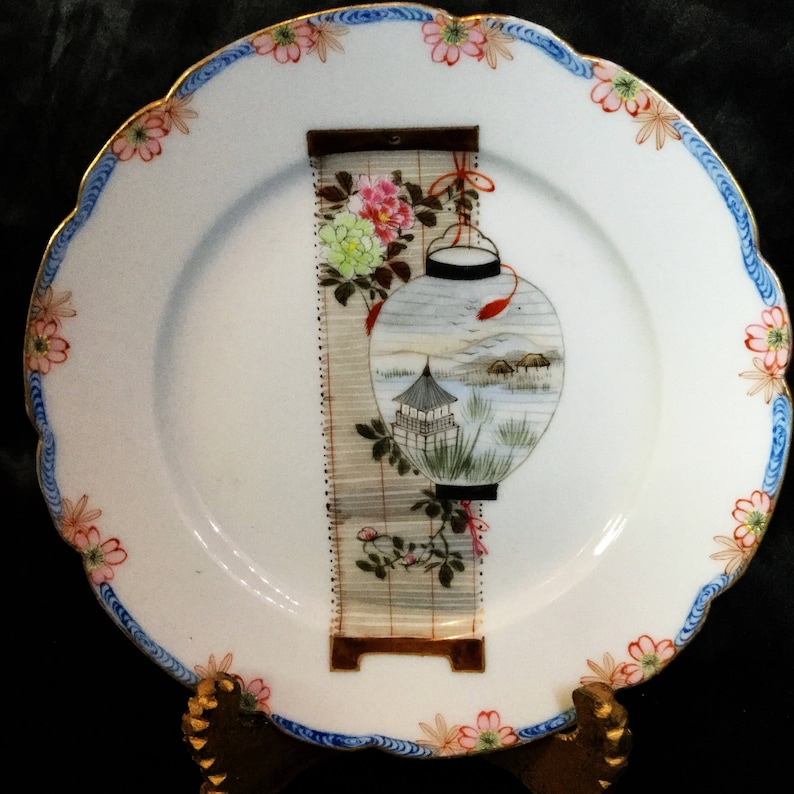 May include: White decorative plate with a scalloped edge, featuring a Japanese-style design. The plate has a blue and gold rim with floral accents. The central image depicts a lantern and a landscape scene with a pagoda.