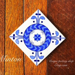May include: A white ceramic tile with a blue and white geometric design. The tile features a central circle with a star-shaped design and a repeating pattern of blue and white arrows. The tile is surrounded by a border of blue and white floral motifs. The tile is labelled "Minton".