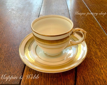 Puede incluir: Una taza de té y un platillo blancos con ribetes dorados. El platillo tiene una banda dorada alrededor del borde y un diseño dorado en el centro. La taza de té tiene una banda dorada alrededor de la parte superior y un diseño dorado alrededor de la parte inferior. El texto "Mappin & Webb" es visible en la esquina inferior izquierda de la imagen. El texto "Unique pottery shop" es visible en la esquina superior derecha de la imagen.