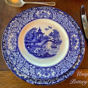 May include: A blue and white porcelain dinner plate with a floral pattern and a central design of a pagoda and bridge. The plate is set on a wooden table with cutlery.