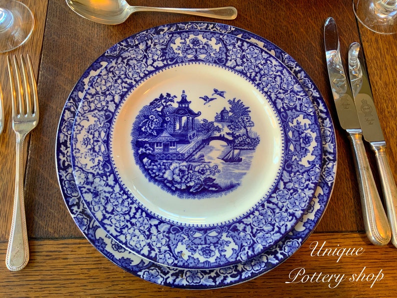 May include: A blue and white porcelain dinner plate with a floral pattern and a central design of a pagoda and bridge. The plate is set on a wooden table with cutlery.