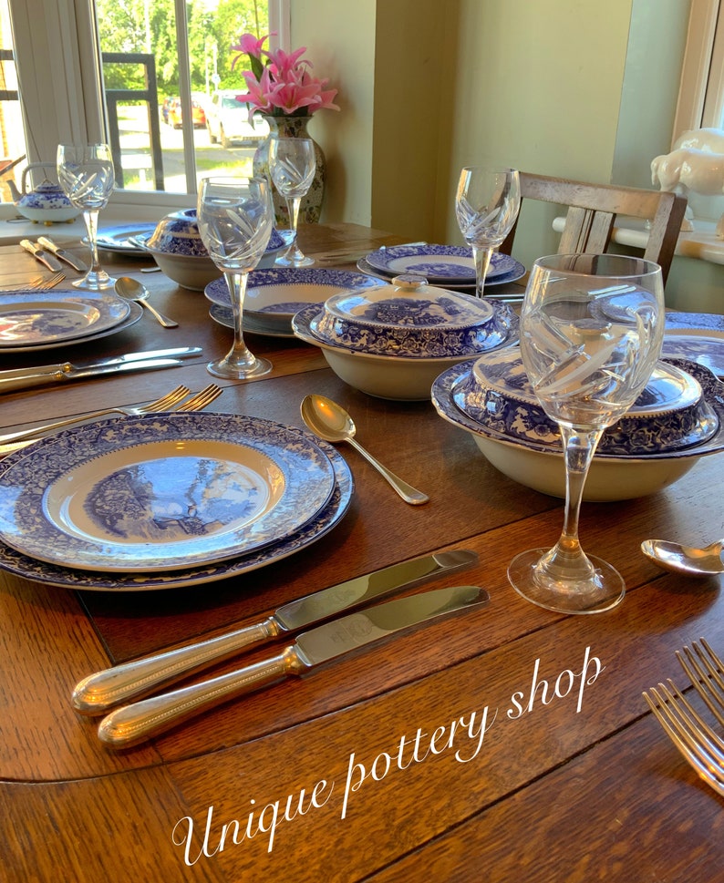 May include: A wooden table set with blue and white patterned china dinnerware, including plates, bowls, and a serving dish. Crystal wine glasses and silver cutlery are also on the table. The text "Unique pottery shop" is written on the table.