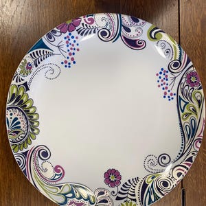 May include: A round white dinner plate featuring a decorative border pattern of swirling paisley shapes, small dots, and stylized flowers in shades of purple, lime green, and dark blue. The center of the plate remains plain white and smooth.