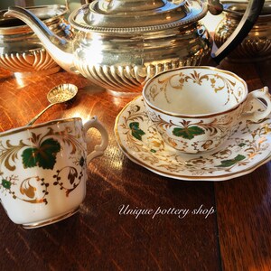 May include: Two teacups and saucers with a gold and green floral pattern. The cups and saucers are on a wooden table with a silver teapot and sugar bowl in the background. The text "Unique pottery shop" is visible in the lower right corner of the image.