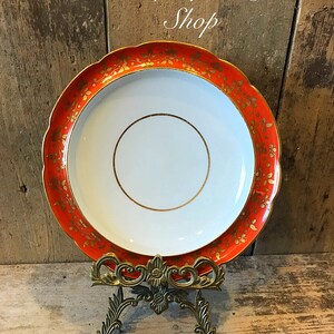 May include: A white ceramic plate with a gold rim and orange floral design. The plate is on a gold metal stand. Unique Pottery Shop.