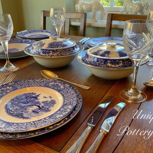 May include: A table setting featuring blue and white patterned china plates, bowls, and serving dishes. The table is made of dark wood and has a silver knife and fork on each place setting. The text "Unique Pottery Shop" is visible in the lower right corner of the image.