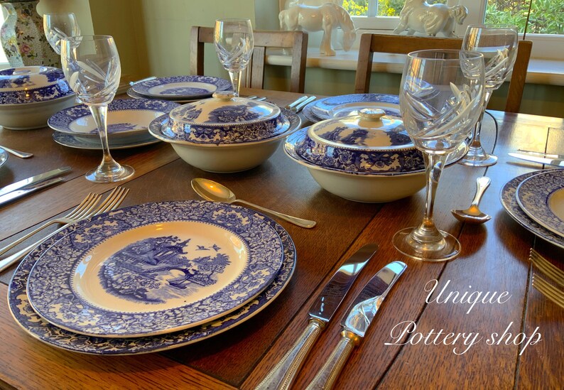 May include: A table setting featuring blue and white patterned china plates, bowls, and serving dishes. The table is made of dark wood and has a silver knife and fork on each place setting. The text "Unique Pottery Shop" is visible in the lower right corner of the image.