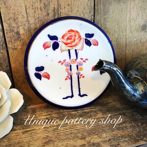 Könnte beinhalten: Ein weißer Keramikteller mit blauem Rand und einem handgemalten Design einer roten und orangen Rose mit grünen Blättern und rosa Blüten. Der Teller liegt auf einer Holzoberfläche.