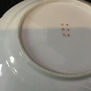 May include: A white ceramic plate with a scalloped edge. The underside features a faded orange inscription in Japanese characters. The plate has a slightly raised rim with a thin, brownish band.