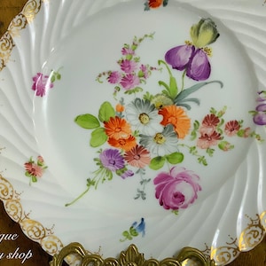 Könnte beinhalten: Ein weißer Porzellanteller mit goldfarbenem Rand und einem Blumendesign. Die Blumen sind in Rosa-, Lila-, Orange- und Gelbtönen bemalt.