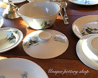 Op de afbeelding: Een set witte keramische borden en kommen met een gouden rand en een eendenontwerp. De borden en kommen zijn op een houten tafel gerangschikt. De tekst "Unique pottery shop Etsy.com" is zichtbaar in de rechterbenedenhoek van de afbeelding.