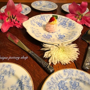 Puede incluir: Un juego de platos de porcelana azul y blanco con un diseño floral. Los platos están dispuestos sobre una mesa de madera con un cuchillo de pastel plateado y una flor blanca. El texto "Unique pottery shop" es visible en la imagen.