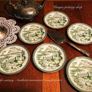 May include: Six green and white transferware plates with a floral and landscape design. The plates are arranged in a circle on a wooden surface. A silver teapot and a silver cake server are also visible in the image. The text "Unique pottery shop" and "19th century Aesthetic movement transferware" are visible in the image.