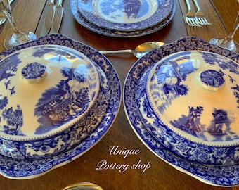 Op de afbeelding: Twee blauwe en witte porseleinen serveerschalen met deksels. De schalen hebben een bloemmotief en een scène met een huis en bomen. De deksels hebben een knop bovenop.
