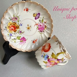 Könnte beinhalten: Ein weißes Porzellan-Teetassen- und Untertassen-Set mit handbemalten Blumendesigns in Rosa-, Gelb-, Orange- und Blautönen. Die Untertasse hat einen gewellten Rand und eine goldene Verzierung. Die Tasse hat ein zartes Blumenmuster und einen goldenen Henkel.