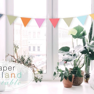 Può includere: Una ghirlanda di carta colorata con bandierine in tonalità di blu, verde, rosa, arancione e giallo è appesa davanti a una finestra. Il testo "Paper Garland printable" è scritto in un ovale bianco su uno sfondo grigio chiaro.