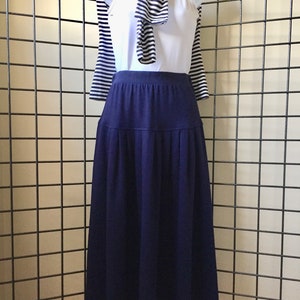 May include: A white vest top, navy blue skirt, and striped sweater draped over a mannequin. The skirt is a solid navy blue, and the sweater has navy and white stripes. The outfit is displayed against a grid background.