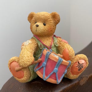 May include: A small, brown teddy bear figurine wearing overalls and holding a red and blue drum with a wooden drumstick.