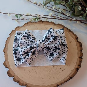 May include: A white baby headband with a large bow featuring a black and white pattern of bows, flowers, and soccer balls. The headband is displayed on a wooden slice with a white background and floral accents.