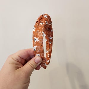May include: An orange headband with a white ghost pattern. The headband is tied in a knot at the top.