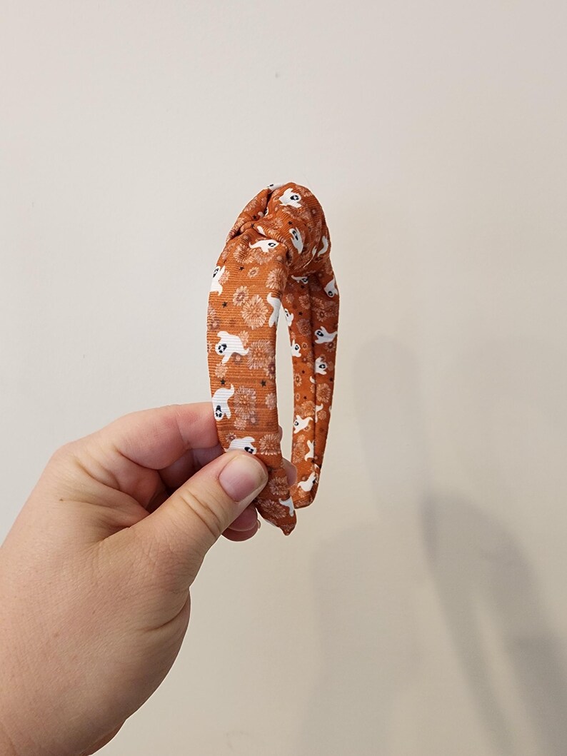 May include: An orange headband with a white ghost pattern. The headband is tied in a knot at the top.