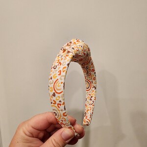 May include: A white headband with a brown, orange, and yellow abstract pattern.