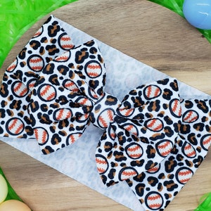 May include: A white headband with a large bow featuring a baseball and leopard print design. The baseballs have red stitching and are set against a white background. The leopard spots are black and brown.