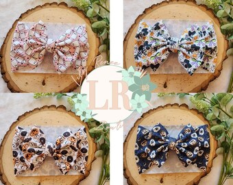 May include: Four different Halloween-themed hair bows. The bows are made of fabric with various patterns, including ghosts, pumpkins, and bats. The bows are displayed on wooden rounds.